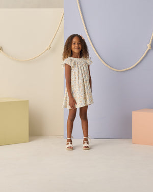 Indoors, a young girl wears the Girls' Lucia Dress by Rylee & Cru, a charming blue floral summer dress, paired with white sandals; pastel blocks and rope decorations adorn the background.