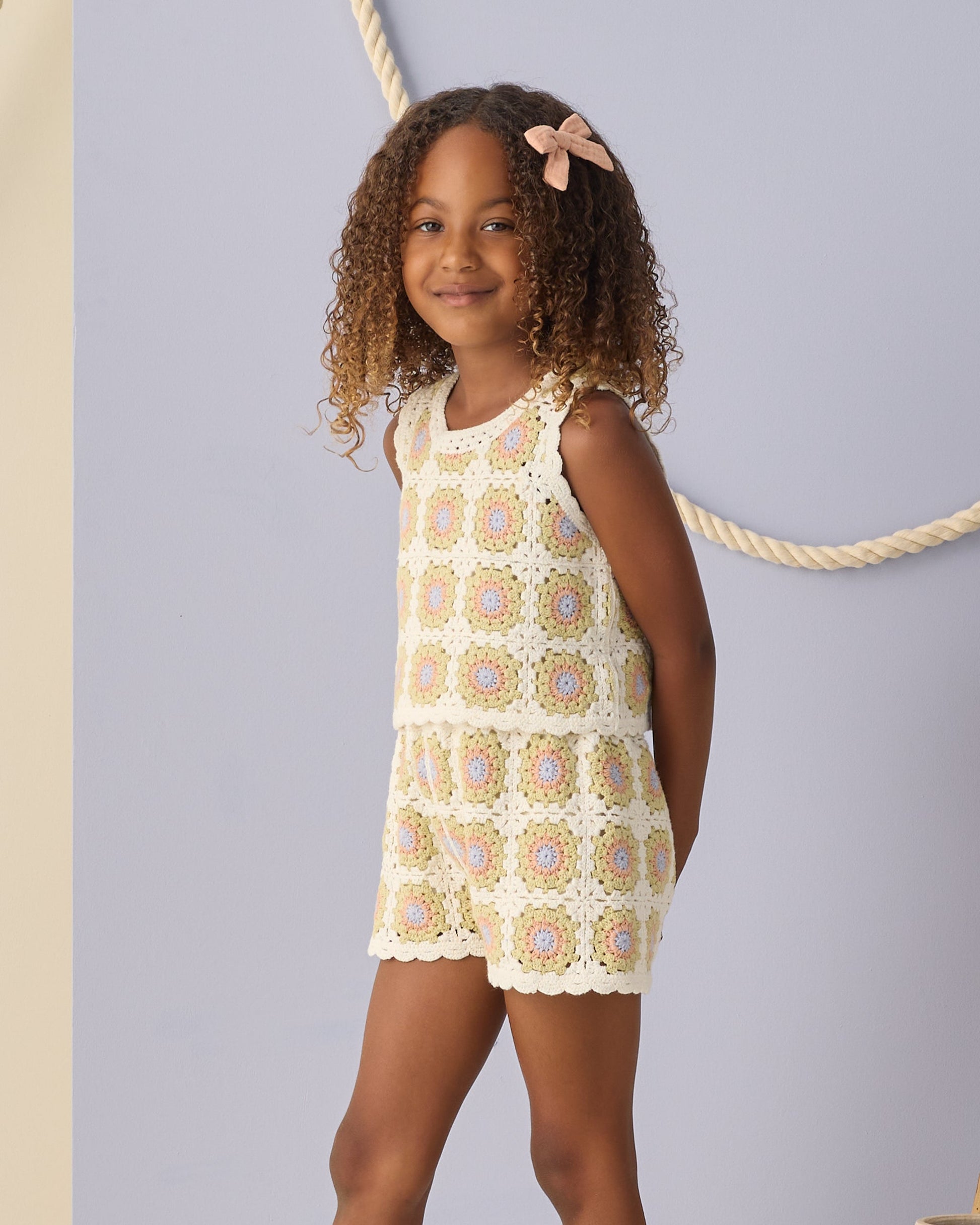 A young girl with curly hair wears the Rylee & Cru Girls' Crochet Tank Set, featuring ruffled trim, standing before a light blue wall with a rope decoration.