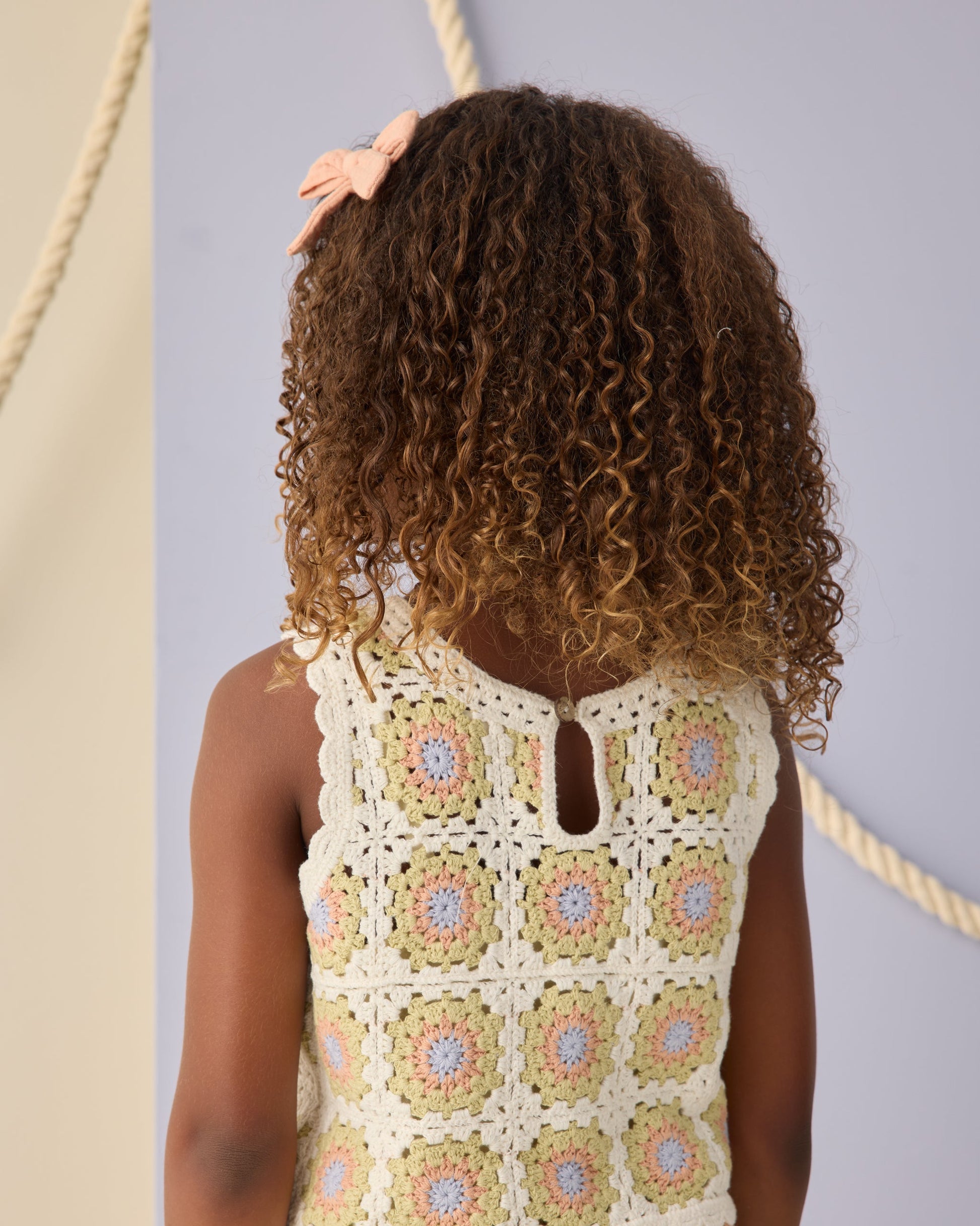 A child with curly hair and a pink bow is seen from behind, wearing the Rylee & Cru Girls' Crochet Tank Set featuring a pastel floral square pattern and delicate ruffled trim.