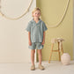 Indoors, a child wears the Rylee & Cru Boys' Lapel Collar Shirt in sea green with matching shorts and beige shoes. A stool and decorative vase appear in the background.