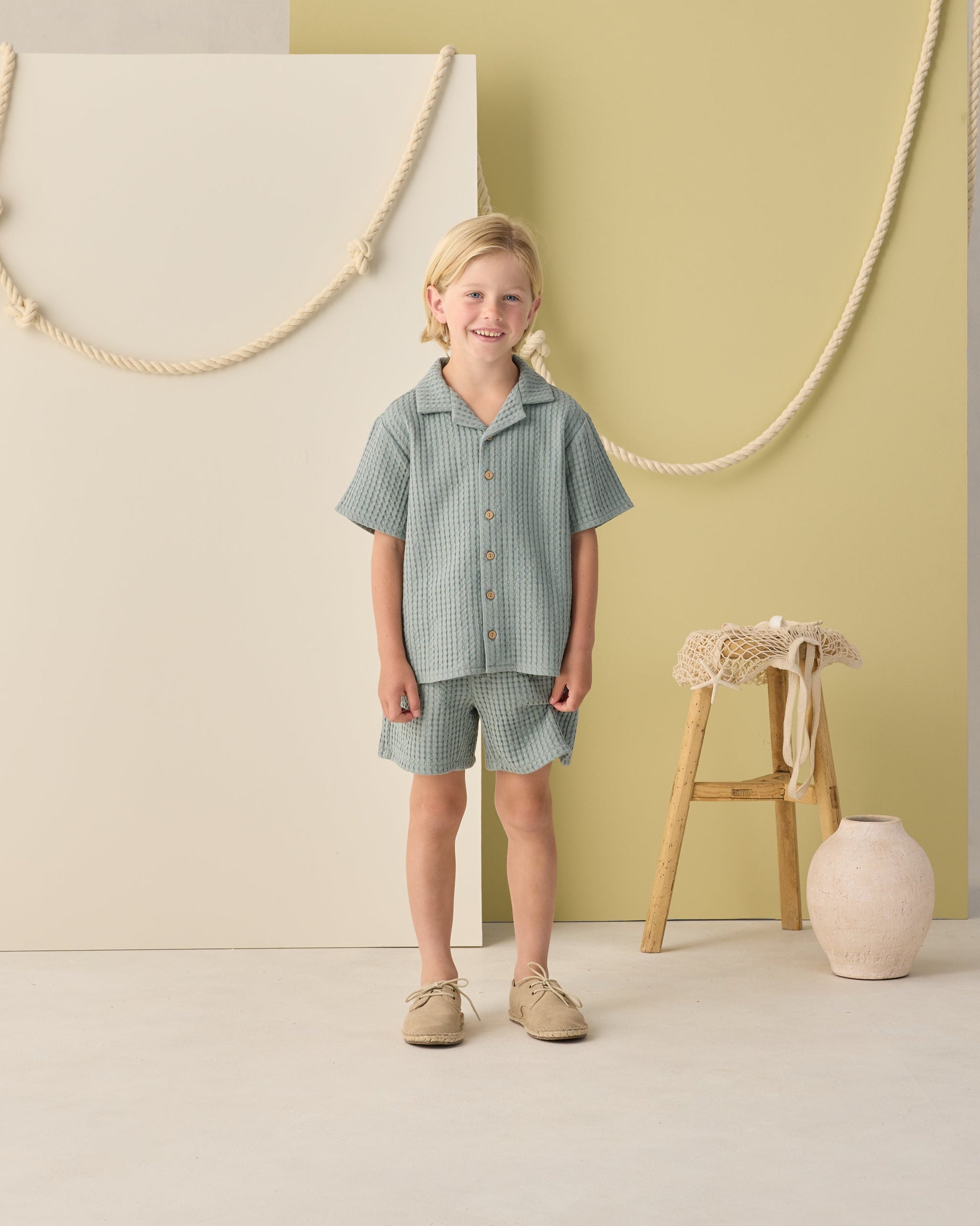 Indoors, a child wears the Rylee & Cru Boys' Lapel Collar Shirt in sea green with matching shorts and beige shoes. A stool and decorative vase appear in the background.