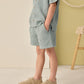 A child stands with hands in pockets, wearing a sea green textured shirt and Rylee & Cru Boys' Relaxed Short, paired with beige shoes. The neutral background includes a rope and wooden stool.