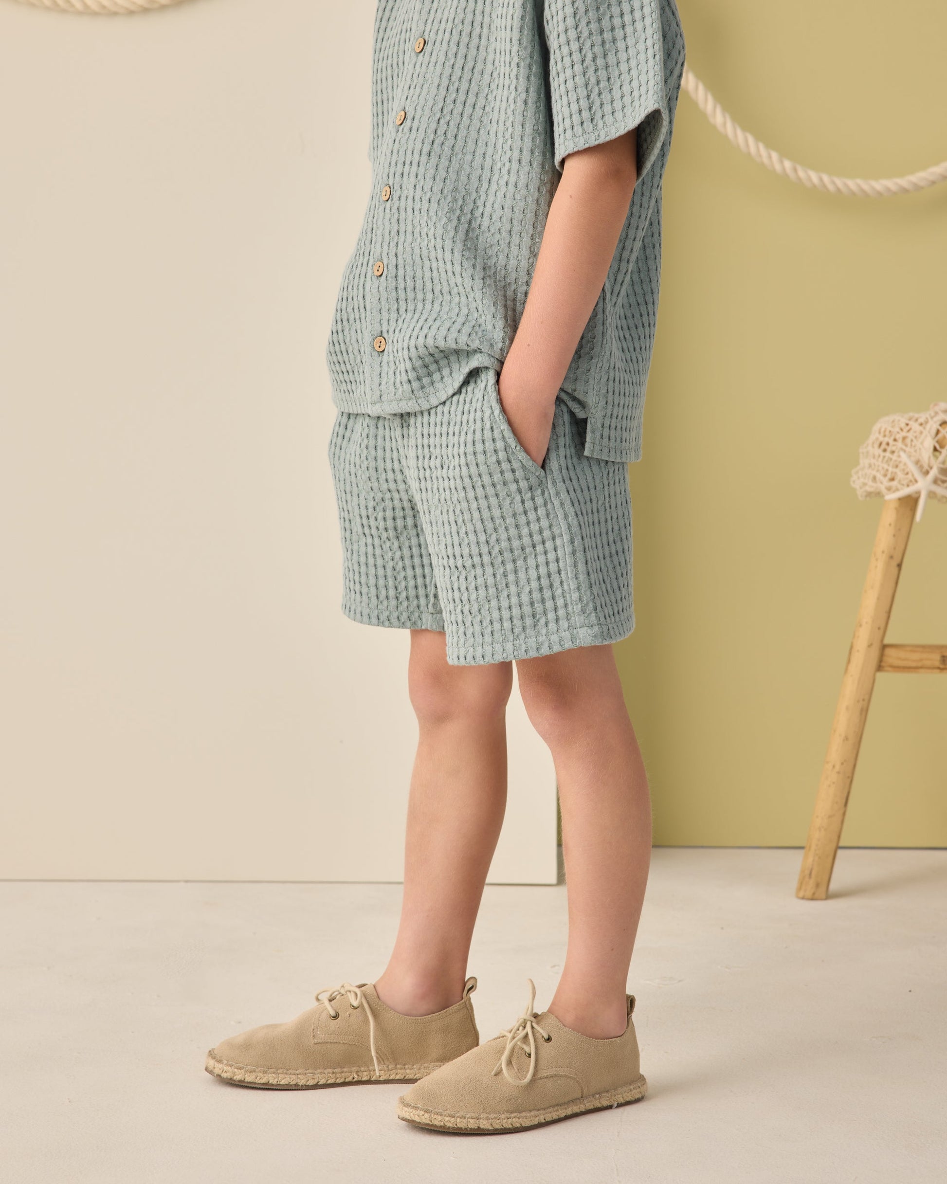 A child stands with hands in pockets, wearing a sea green textured shirt and Rylee & Cru Boys' Relaxed Short, paired with beige shoes. The neutral background includes a rope and wooden stool.