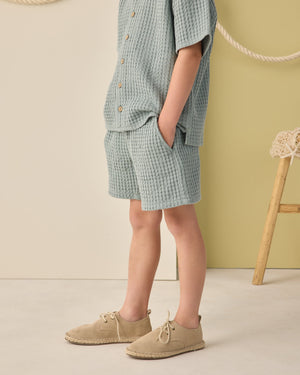 A child stands with hands in pockets, wearing a sea green textured shirt and Rylee & Cru Boys' Relaxed Short, paired with beige shoes. The neutral background includes a rope and wooden stool.
