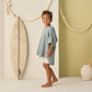 A young child wears the Rylee & Cru Kids' Beach Poncho in sea green, standing barefoot indoors beside a surfboard and large rope on a light backdrop.