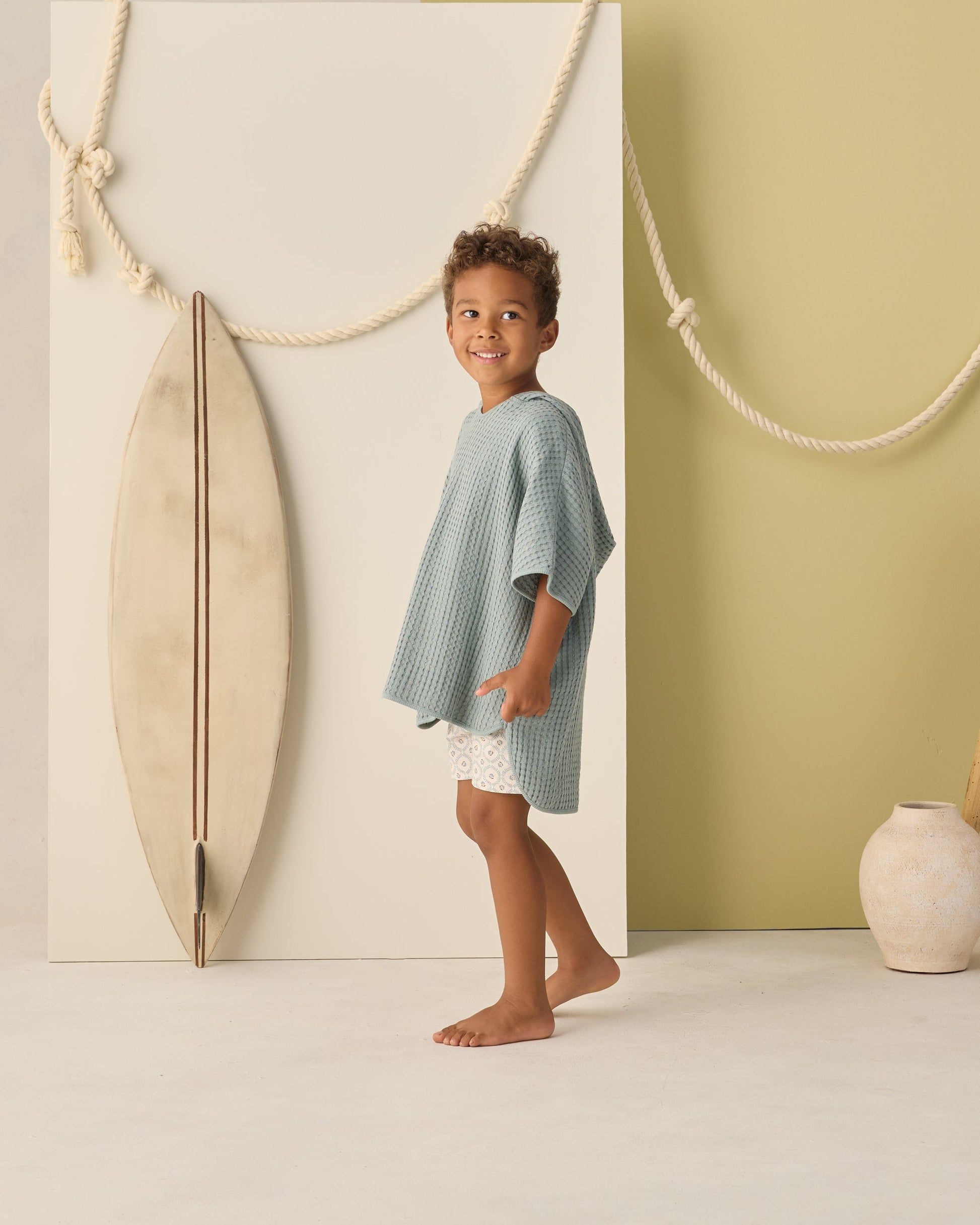 A young child wears the Rylee & Cru Kids' Beach Poncho in sea green, standing barefoot indoors beside a surfboard and large rope on a light backdrop.