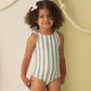 A young girl with curly hair wears the Rylee & Cru Girls' Scrunch One-Piece in light blue and white stripes, standing before a wall decorated with hanging ropes.