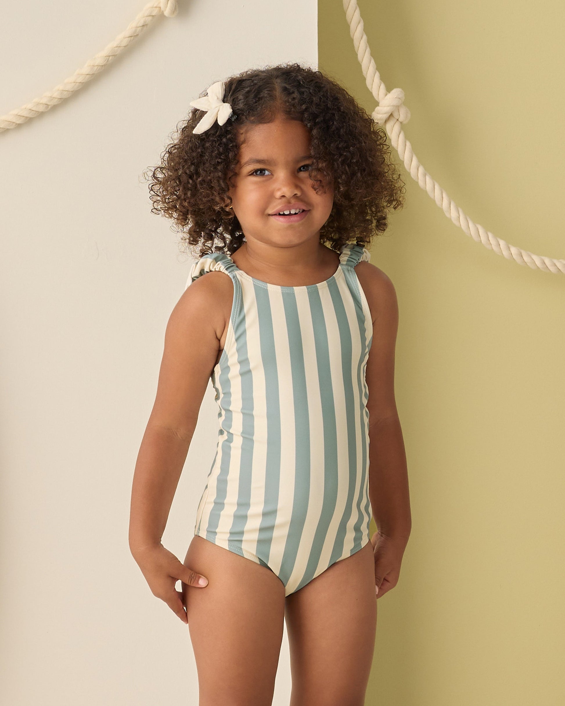 A young girl with curly hair wears the Rylee & Cru Girls' Scrunch One-Piece in light blue and white stripes, standing before a wall decorated with hanging ropes.