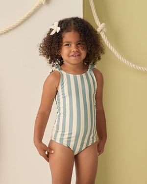 A young girl with curly hair wears the Rylee & Cru Girls' Scrunch One-Piece in light blue and white stripes, standing before a wall decorated with hanging ropes.