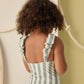 A child with curly hair, seen from behind, wears the Rylee & Cru Girls' Scrunch One-Piece featuring ruffled straps and light blue and white stripes while standing by a rope-adorned wall.