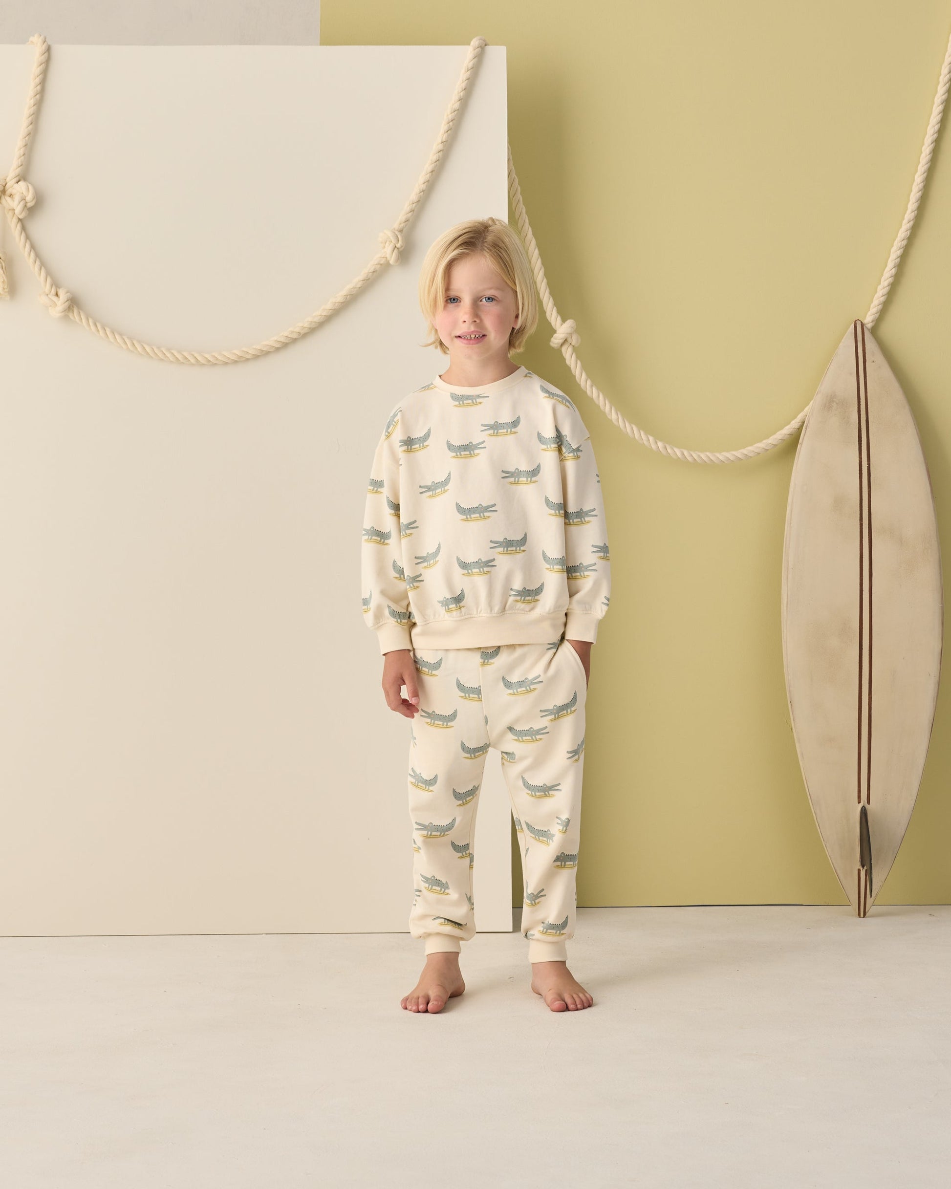 A child stands barefoot indoors wearing the Rylee & Cru Baby Boys' and Boys' Relaxed Sweatshirt with an alligator print, next to a surfboard and decorative ropes on a pale green and white background.