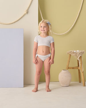 A young girl stands indoors wearing the Rylee & Cru Girls' Cropped Rashguard Set, surrounded by neutral and pastel decor featuring a rope, stool, basket, and vase in the background.