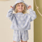 A young girl smiles in front of a beige wall with rope decor, wearing the Rylee & Cru Baby Girls' and Girls' Karli Set—a periwinkle daisy print summer outfit with matching shorts and hat.