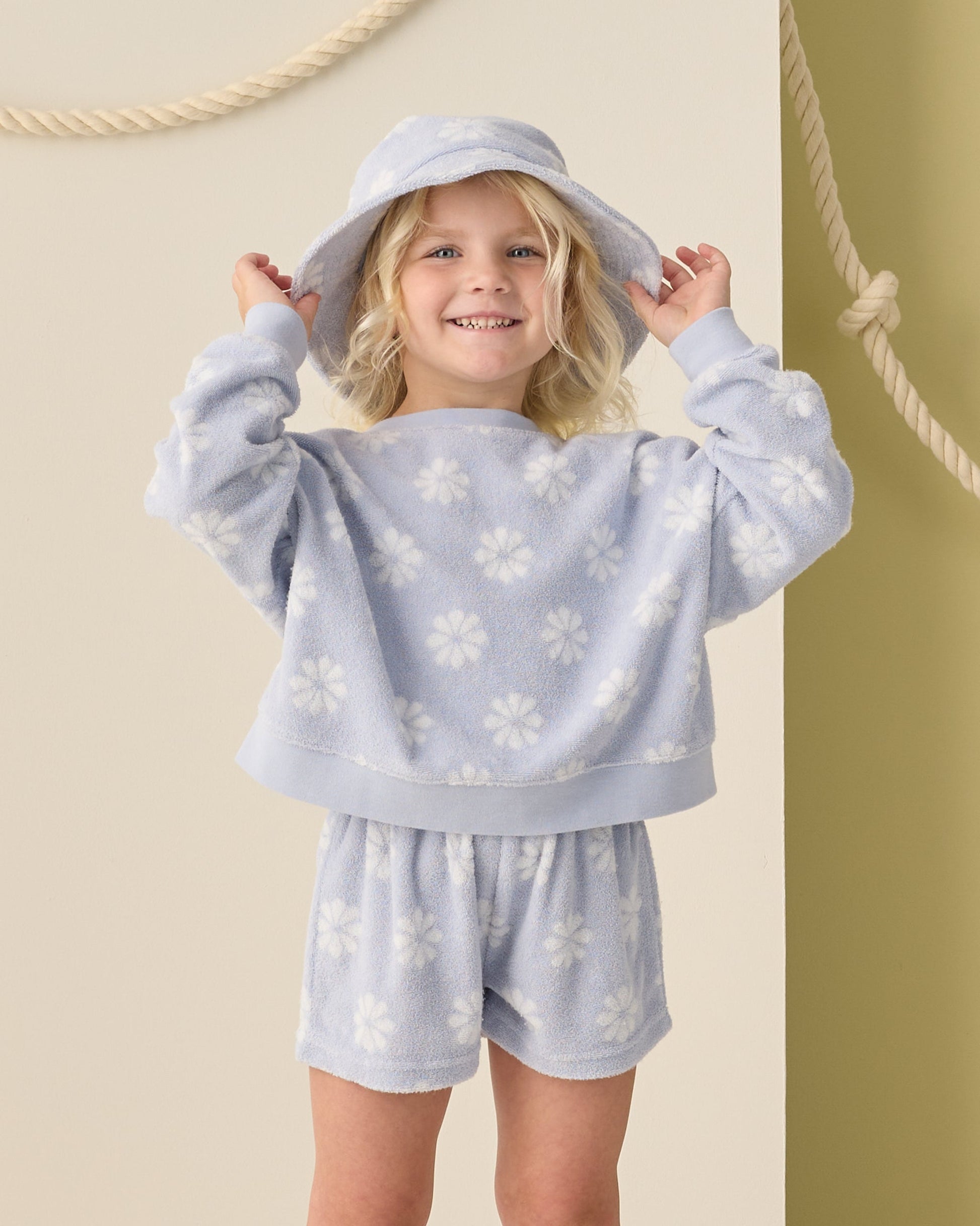 A young girl smiles in front of a beige wall with rope decor, wearing the Rylee & Cru Baby Girls' and Girls' Karli Set—a periwinkle daisy print summer outfit with matching shorts and hat.