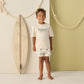 A young boy stands indoors beside a surfboard, wearing Rylee & Cru Baby Boys' and Boys' Swim Trunk with an elastic drawstring and mesh liner; rope decor hangs on the wall behind him.