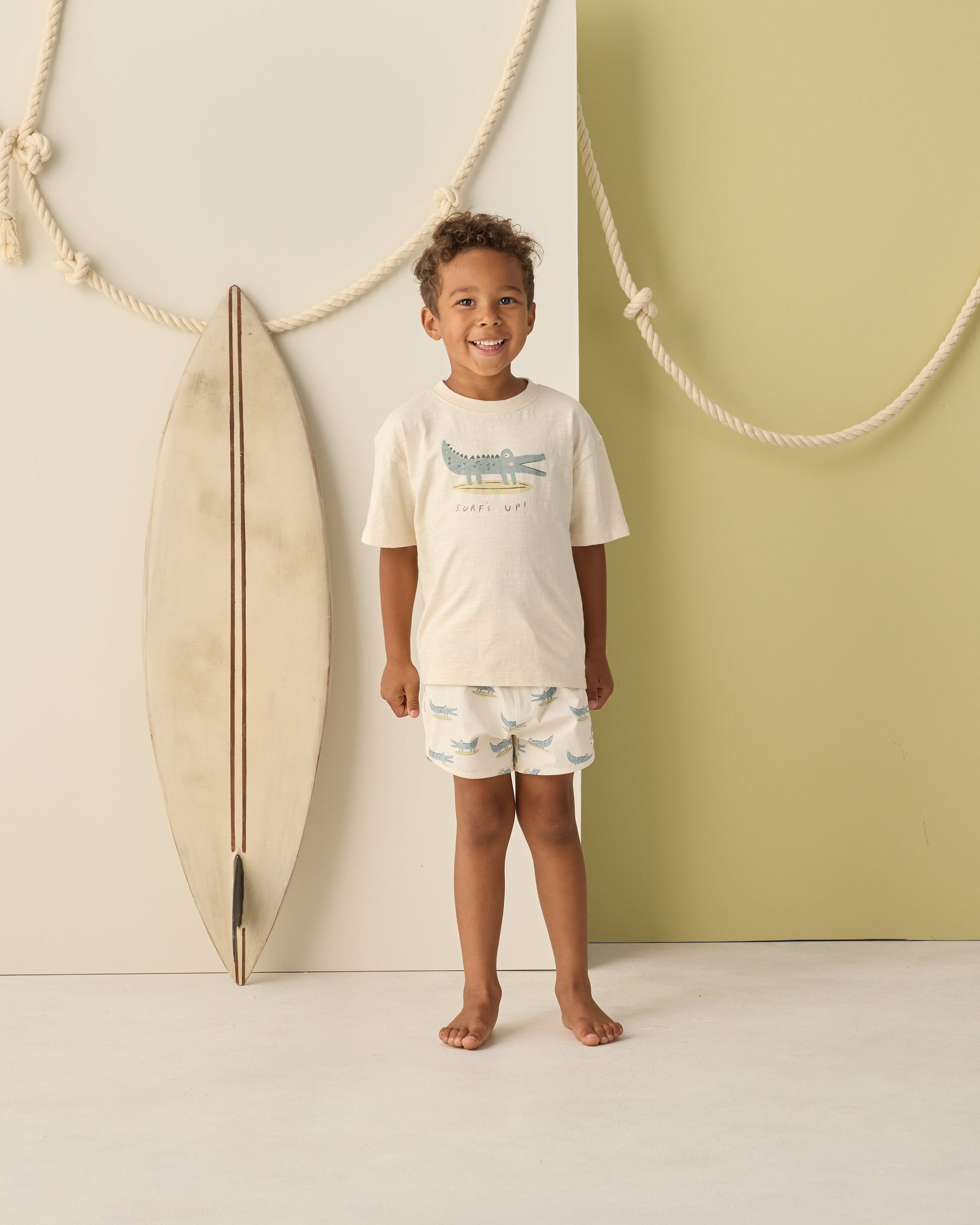 A young boy stands indoors beside a surfboard, wearing Rylee & Cru Baby Boys' and Boys' Swim Trunk with an elastic drawstring and mesh liner; rope decor hangs on the wall behind him.