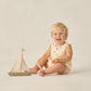 A smiling blonde toddler sits on the floor in a sea green Rylee & Cru Baby Boys' Beau Romper, with a small wooden sailboat in front—a charming summer outfit for playful days.