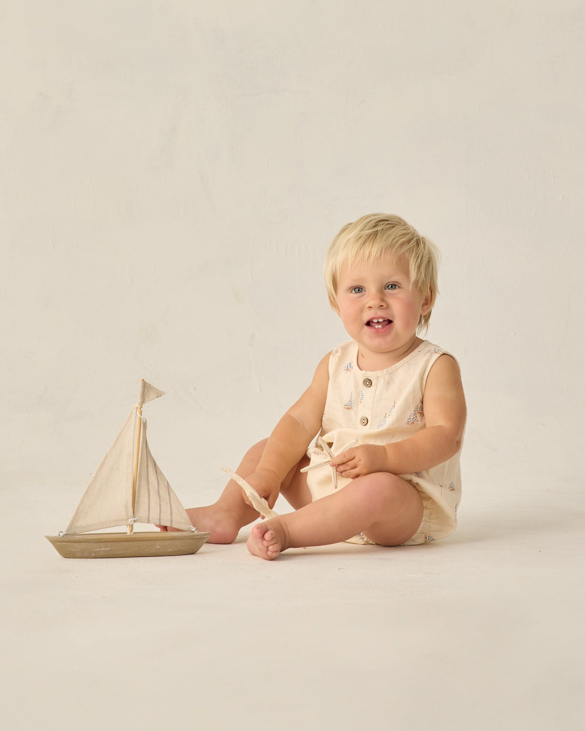 A smiling blonde toddler sits on the floor in a sea green Rylee & Cru Baby Boys' Beau Romper, with a small wooden sailboat in front—a charming summer outfit for playful days.