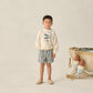 A young boy stands indoors wearing a cream sweater with a fish graphic, Rylee & Cru Baby Boys' and Boys' Dawson Shorts, and beige shoes. Fishing nets and buoys sit to his right.