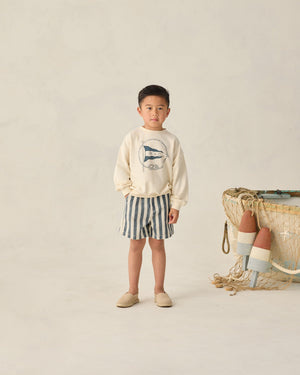 A young boy stands indoors wearing a cream sweater with a fish graphic, Rylee & Cru Baby Boys' and Boys' Dawson Shorts, and beige shoes. Fishing nets and buoys sit to his right.