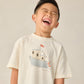 A young boy with short dark hair laughs with his eyes closed, wearing the Rylee & Cru Baby Boys' and Boys' Relaxed Tee in cream, made from soft cotton jersey and featuring a boat illustration.