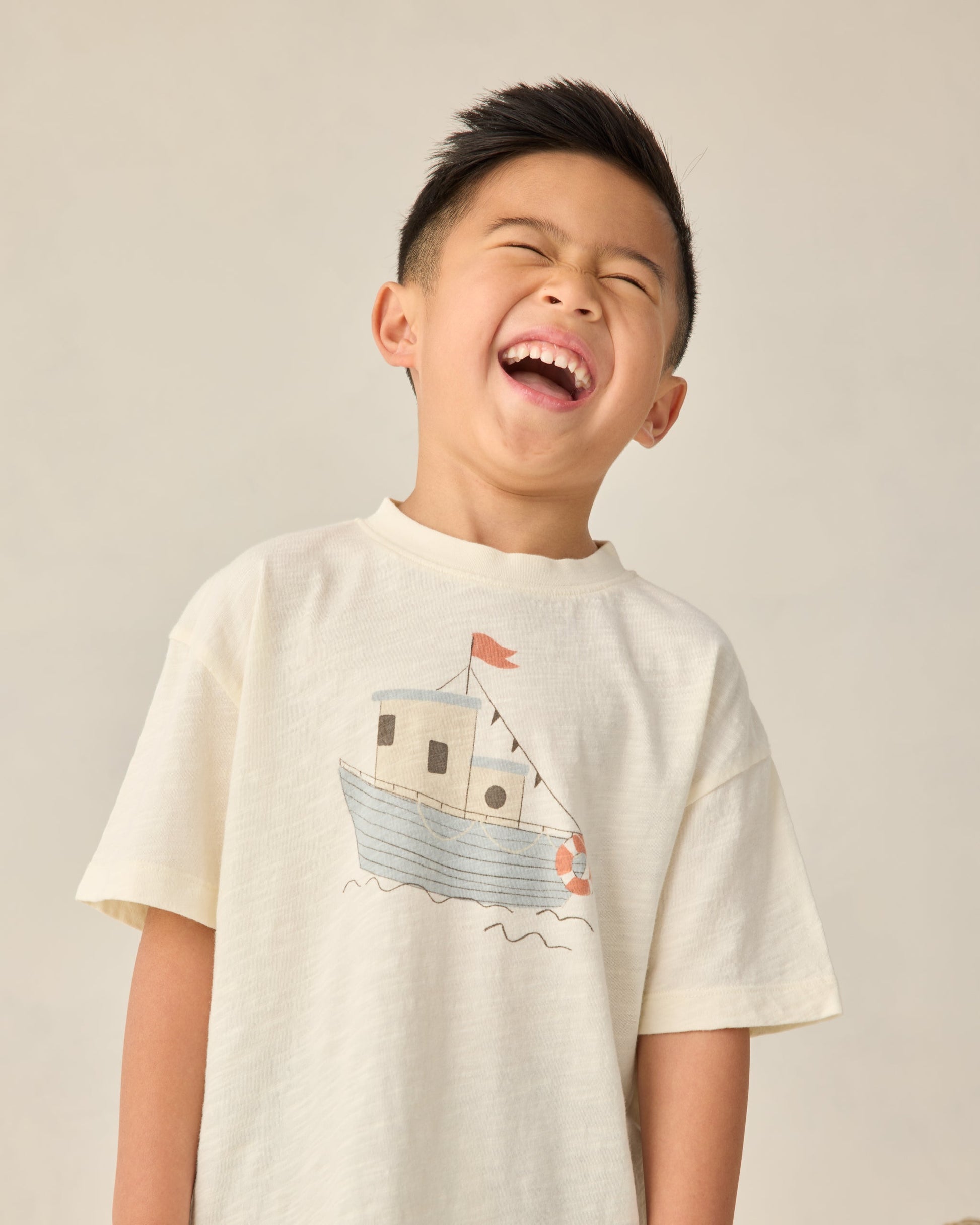 A young boy with short dark hair laughs with his eyes closed, wearing the Rylee & Cru Baby Boys' and Boys' Relaxed Tee in cream, made from soft cotton jersey and featuring a boat illustration.