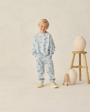 A young boy wearing Rylee & Cru Baby Boys' and Boys' Sweatpant in light blue with a playful sharks print smiles beside a wooden stool holding two ceramic vases, all set against a plain beige background.