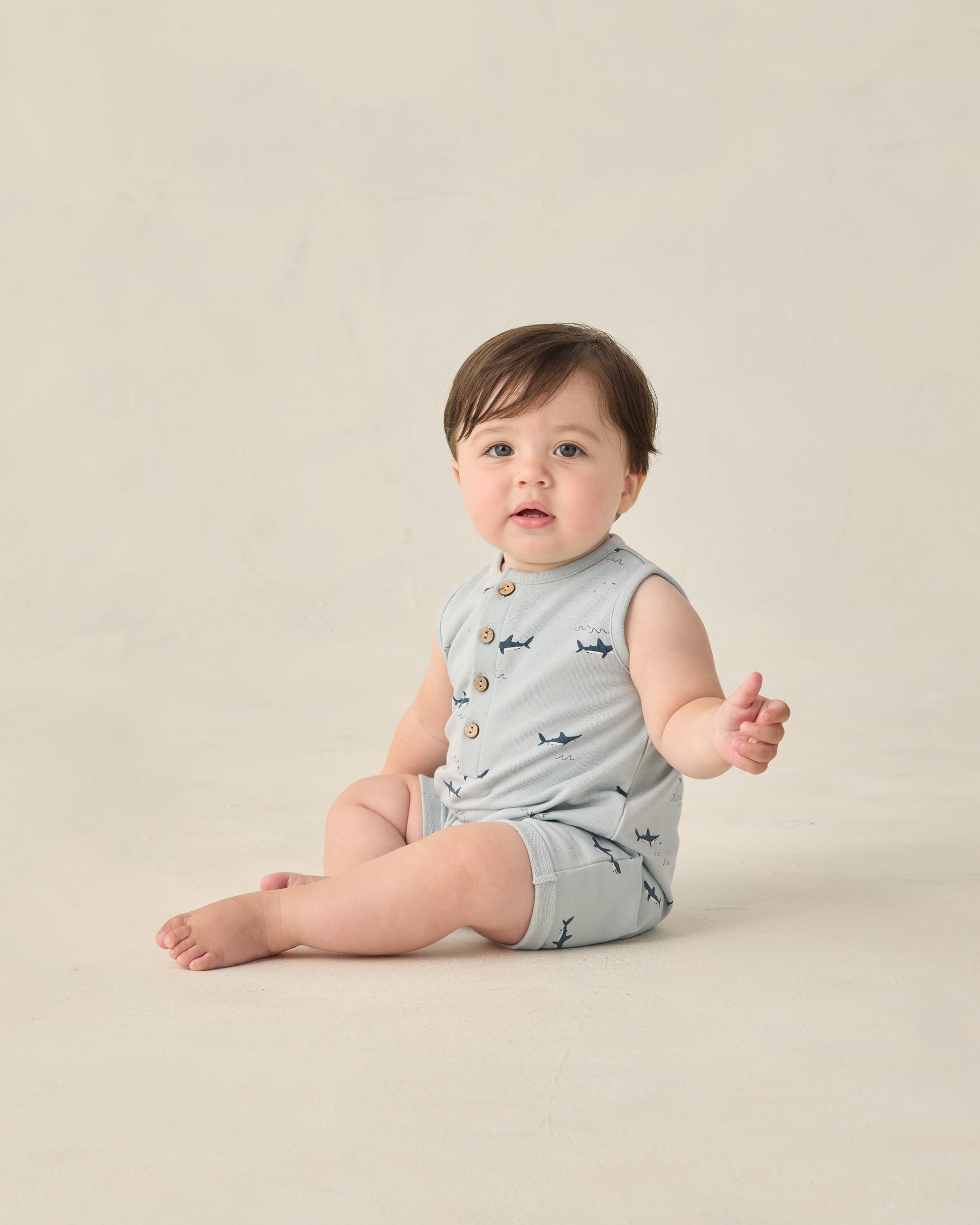 A baby wears the Rylee & Cru Baby Boys' Davis Romper—a sleeveless, light grey outfit with playful sharks print—while sitting on the floor against a neutral background.