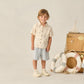 A young child stands indoors by picnic baskets and a model sailboat, wearing the Rylee & Cru Baby Boys' and Boys' Collared Short Sleeve Shirt with comfy shorts and sneakers.