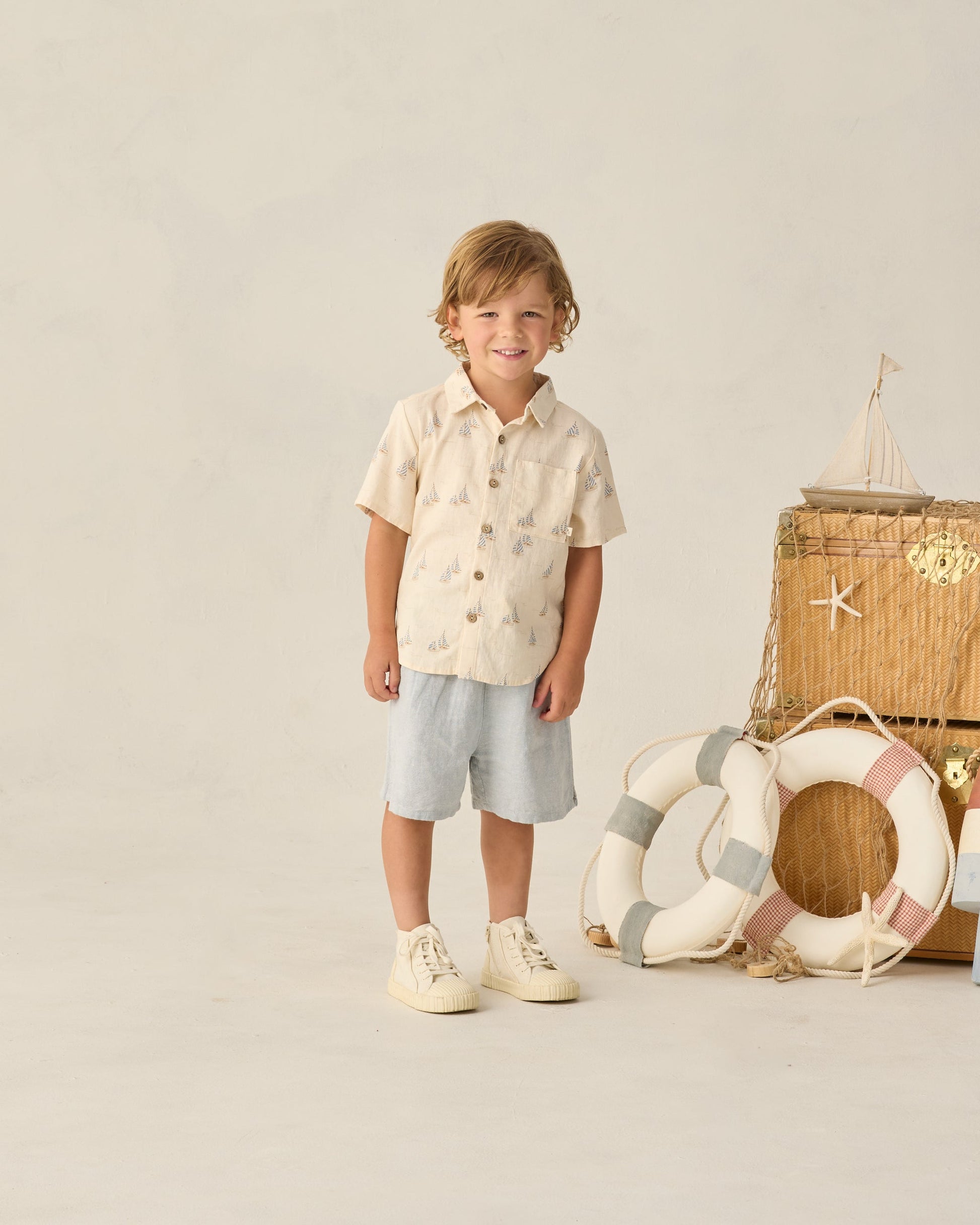 A young boy in Rylee & Cru’s Baby Boys' and Boys' Perry Short with an elastic waistband and short-sleeve shirt stands by suitcases, lifebuoys, and a model sailboat for a playful vintage style against a simple backdrop.