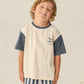 A young boy with wavy hair smiles in a Rylee & Cru Baby Boys' and Boys' Contrast Short Sleeve Tee, featuring cream and navy cotton slub with a small nautical flag graphic, paired with navy-and-white striped shorts.