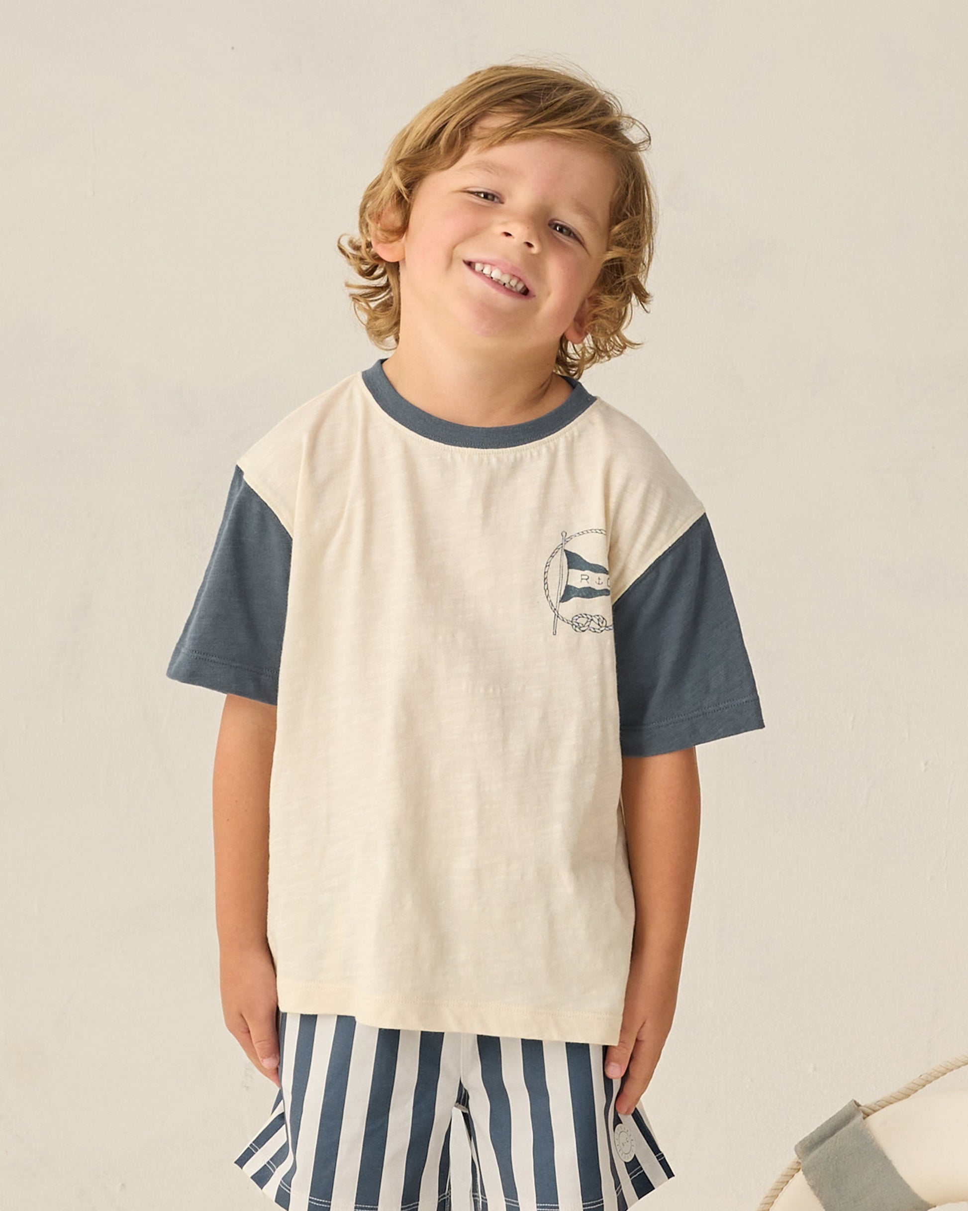 A young boy with wavy hair smiles in a Rylee & Cru Baby Boys' and Boys' Contrast Short Sleeve Tee, featuring cream and navy cotton slub with a small nautical flag graphic, paired with navy-and-white striped shorts.