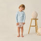 A young boy with light brown hair wears a light blue Rylee & Cru Baby Boys' and Boys' Long Sleeve Rashguard, paired with floral shorts, standing barefoot by a wooden stool with vases and starfish, ready for a beach day.