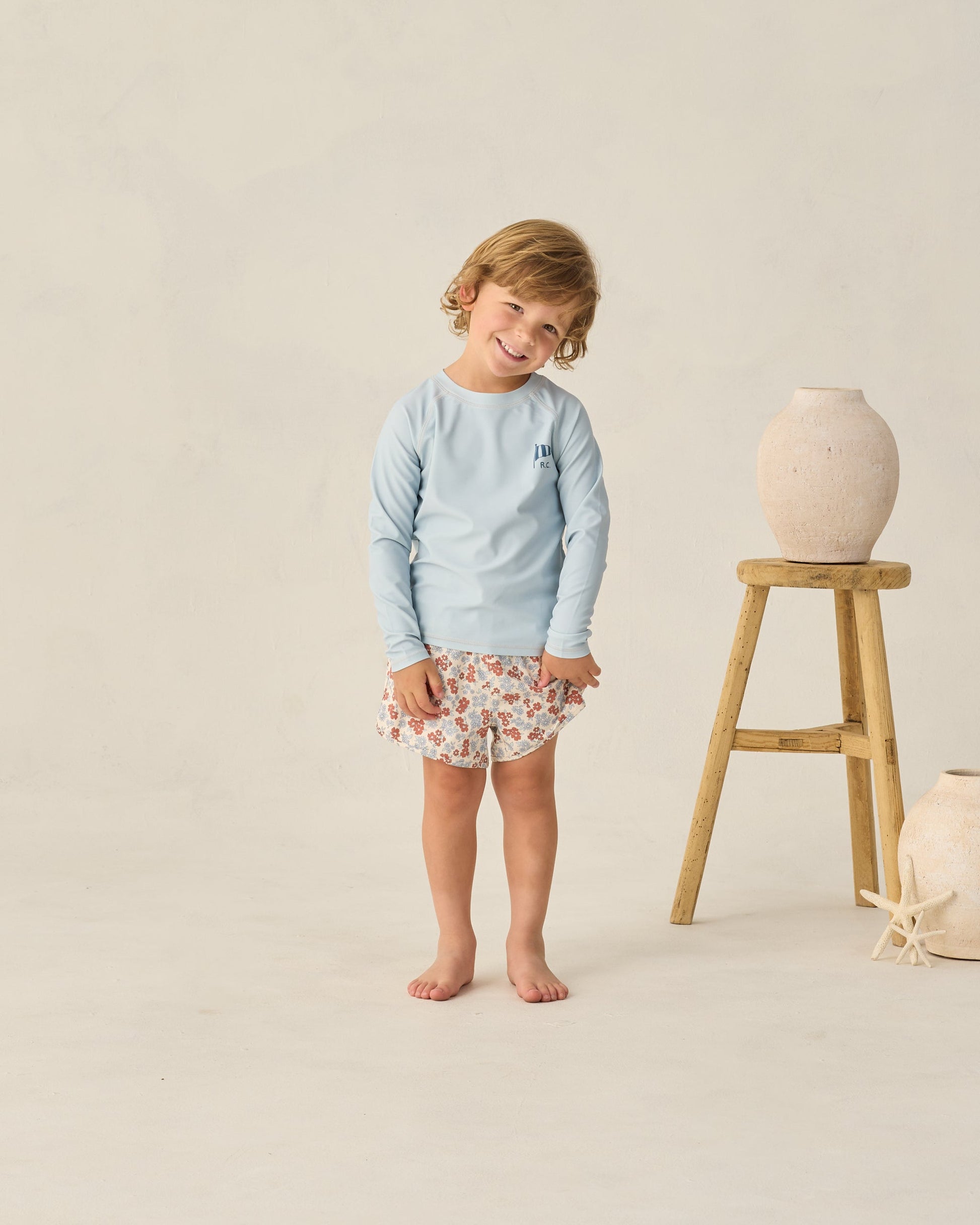 A young boy with light brown hair wears a light blue Rylee & Cru Baby Boys' and Boys' Long Sleeve Rashguard, paired with floral shorts, standing barefoot by a wooden stool with vases and starfish, ready for a beach day.