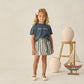 A young girl stands indoors surrounded by ceramic vases and a wooden sailboat, wearing a Rylee & Cru Baby Girls’ and Girls’ Boxy Tee in navy with “Beach Babe” text, paired with a striped skirt.