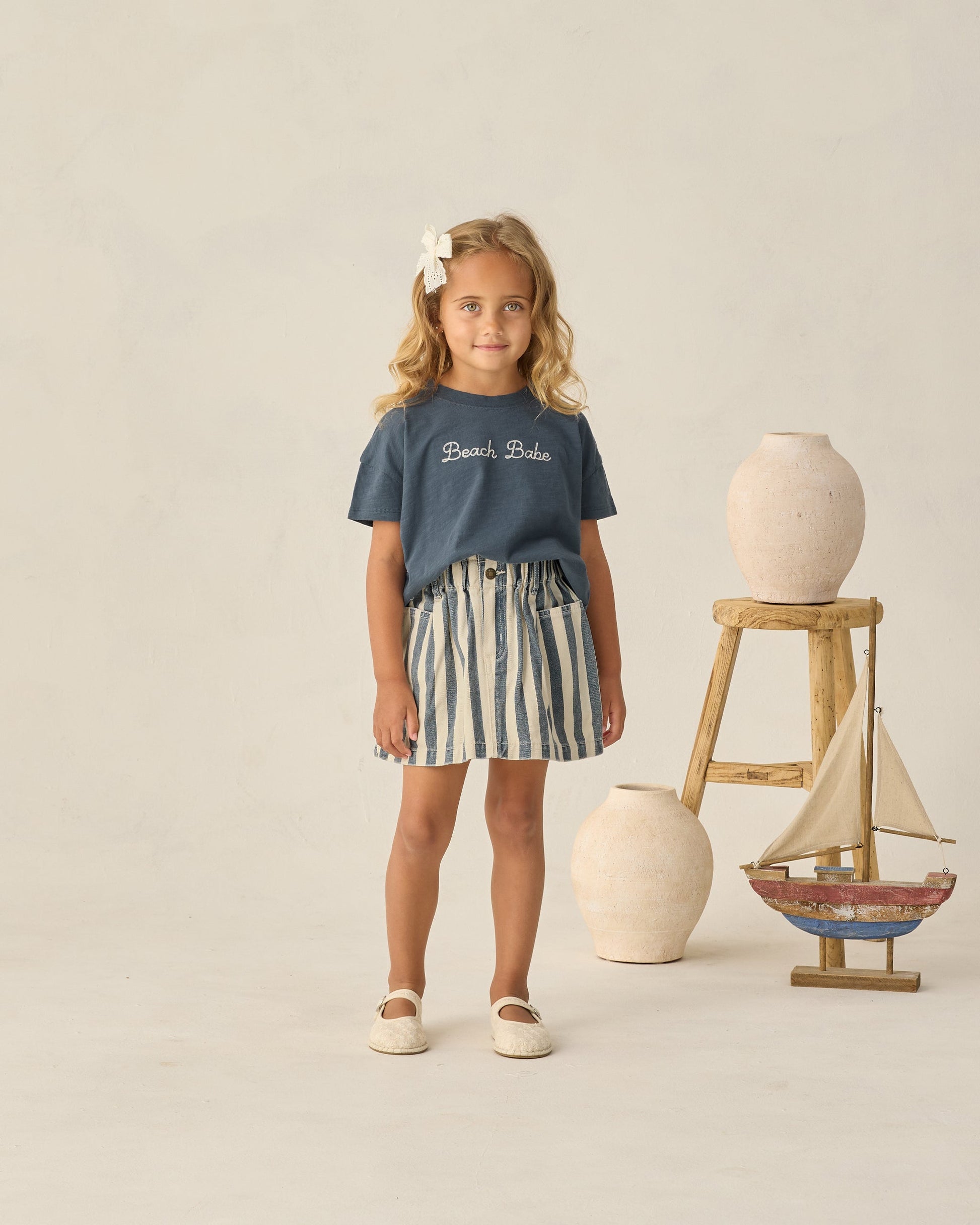 A young girl stands indoors surrounded by ceramic vases and a wooden sailboat, wearing a Rylee & Cru Baby Girls’ and Girls’ Boxy Tee in navy with “Beach Babe” text, paired with a striped skirt.