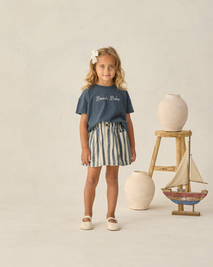 A young girl stands indoors surrounded by ceramic vases and a wooden sailboat, wearing a Rylee & Cru Baby Girls’ and Girls’ Boxy Tee in navy with “Beach Babe” text, paired with a striped skirt.