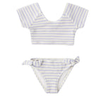 The Rylee & Cru Girls' Cropped Rashguard Set includes a short-sleeve crop top and matching bottoms with light blue and white stripes, side-tie detail, and UPF 50+ protection for stylish, sun-safe swimwear.