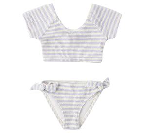 The Rylee & Cru Girls' Cropped Rashguard Set includes a short-sleeve crop top and matching bottoms with light blue and white stripes, side-tie detail, and UPF 50+ protection for stylish, sun-safe swimwear.