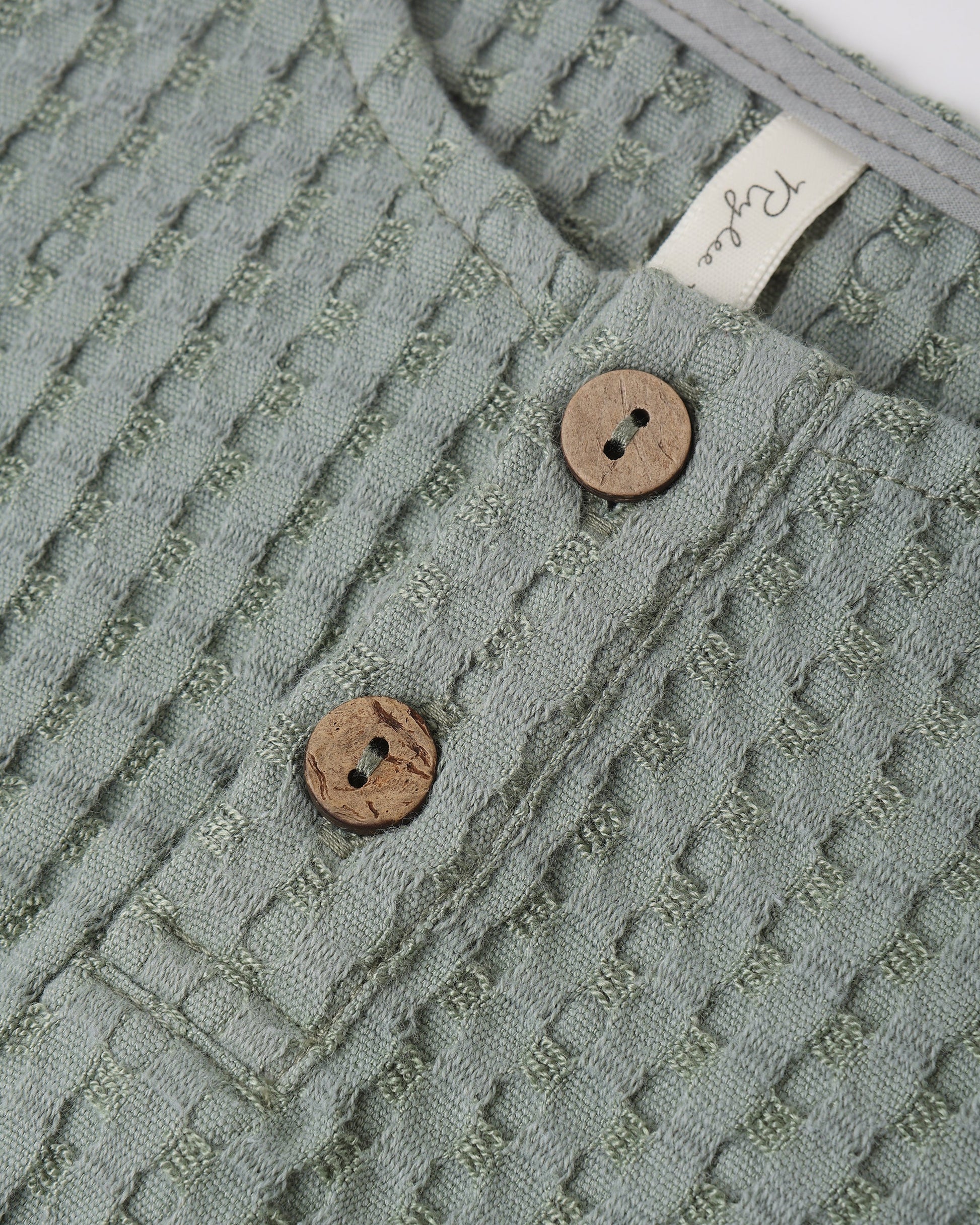 Close-up of Rylee & Cru's Baby Boys' Beau Romper in textured sea green, with two wooden buttons and a white collar tag—a stylish summer outfit.