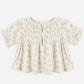 Light-colored, short-sleeved Girls’ Colette Top by Rylee & Cru, featuring a charming floral pattern, smocked detailing at the chest for a flattering fit, ruffled cuffs, and two buttons at the front neckline, displayed on a plain background.