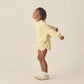 A young child smiles and stretches in the Rylee & Cru Kids' Racer Sweatshirt with matching shorts, white socks, and beige sneakers—an ideal transitional look against a plain background.