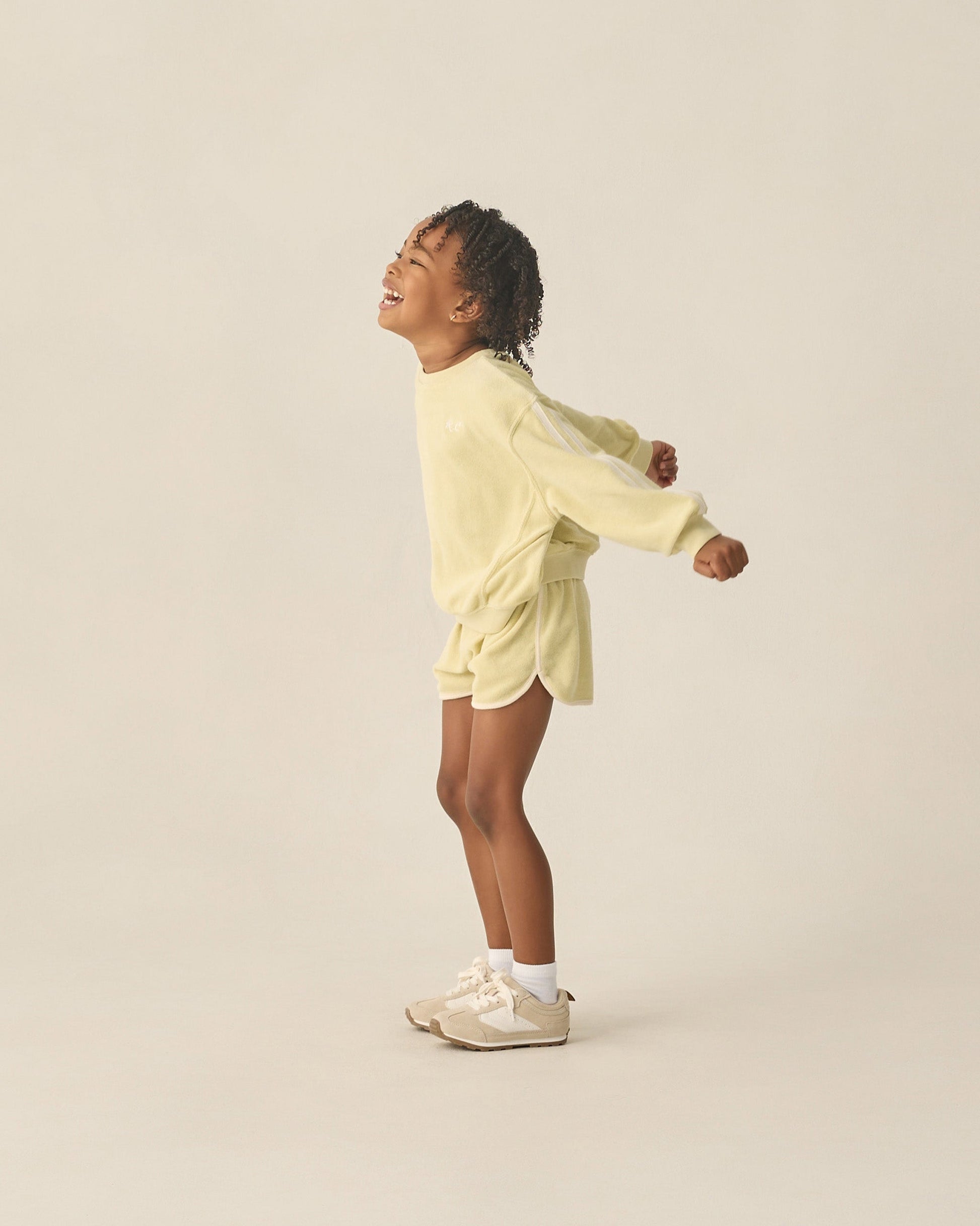 A young child smiles and stretches in the Rylee & Cru Kids' Racer Sweatshirt with matching shorts, white socks, and beige sneakers—an ideal transitional look against a plain background.