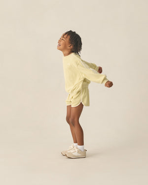 A young child smiles and stretches in the Rylee & Cru Kids' Racer Sweatshirt with matching shorts, white socks, and beige sneakers—an ideal transitional look against a plain background.