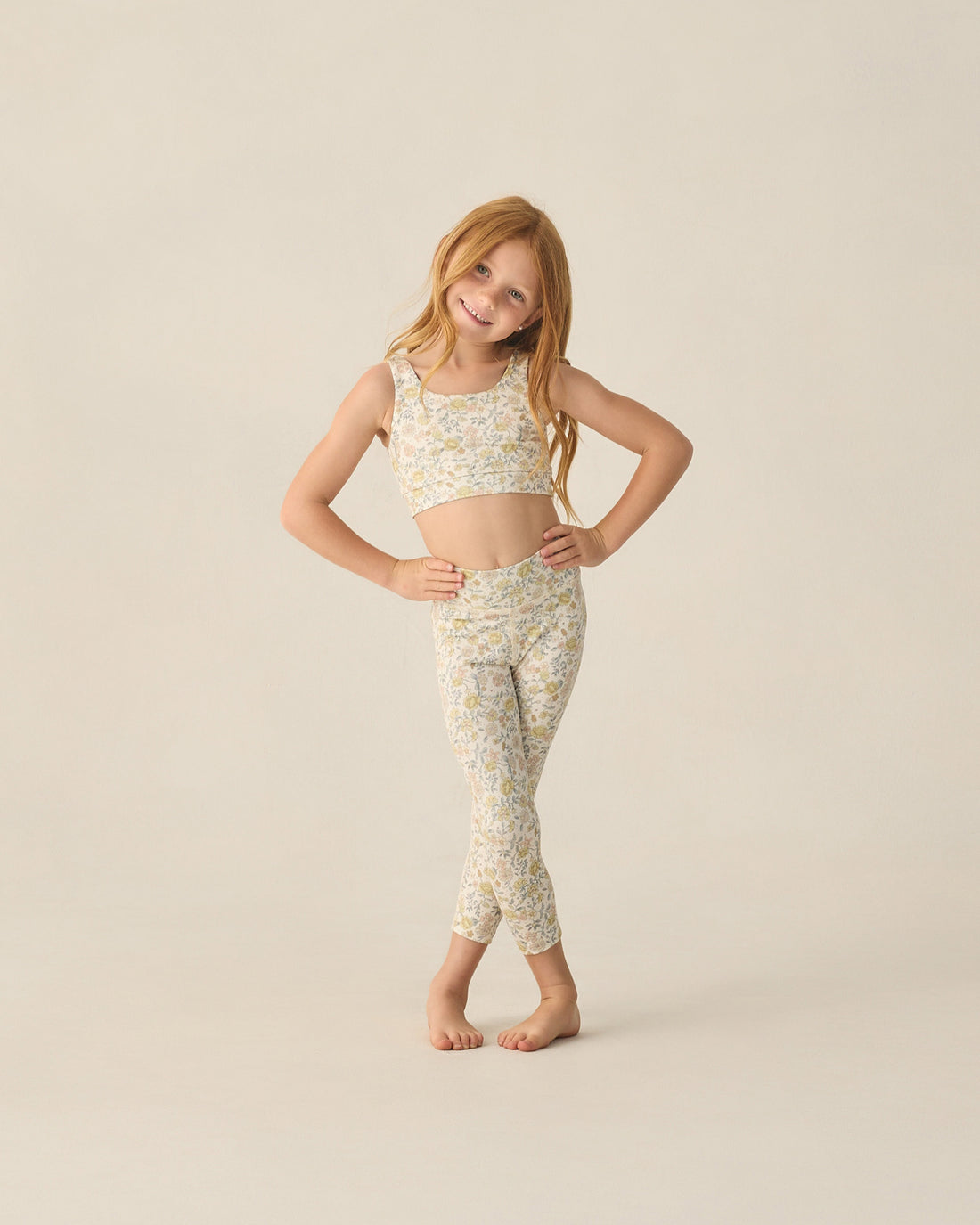 A young girl with long red hair stands barefoot, wearing a Rylee & Cru Girls' Swift Sports Top and matching leggings with a floral print, posing against a plain light background.