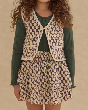 A young girl stands against a plain background in a green ribbed top, floral-patterned vest, and the Girls' Rose Mini Skirt by Rylee & Cru, which features scalloped tiers.