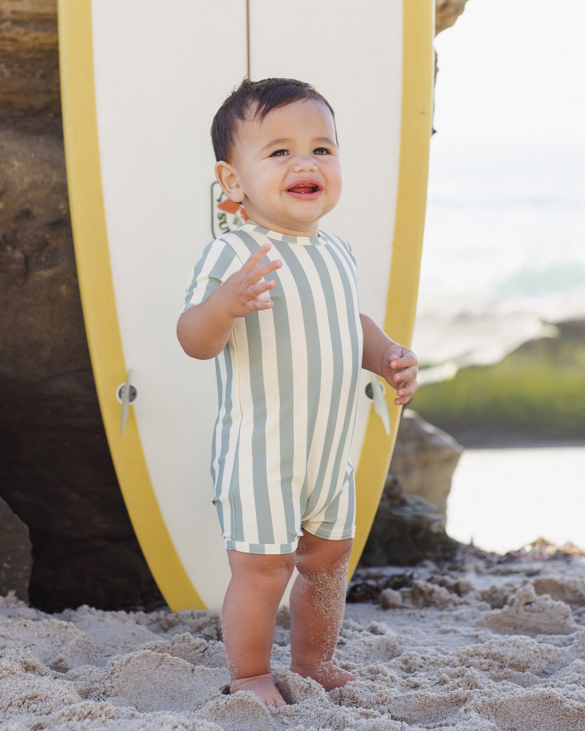 A smiling toddler in a green and white striped Rylee & Cru Baby Boys' Shorty One-Piece stands barefoot on the sand before a yellow and white surfboard, ready for beach fun in stylish swimwear.