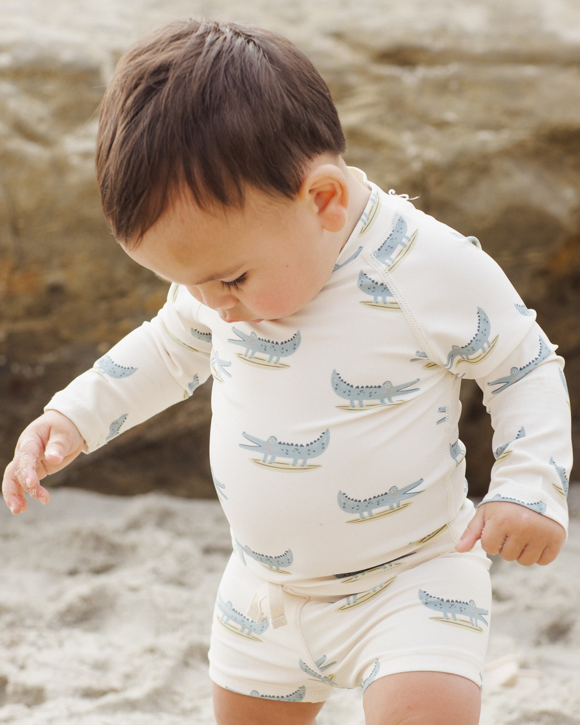 A toddler wears the Rylee & Cru Baby Boys' Rashguard Set with an alligator print, standing on a sandy beach in UPF 50+ swimwear.