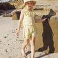 A young child in a yellow outfit and sun hat walks barefoot on a sandy beach near large rocks, wearing comfy Rylee & Cru Girls' Remi Shorts with an elastic waist.