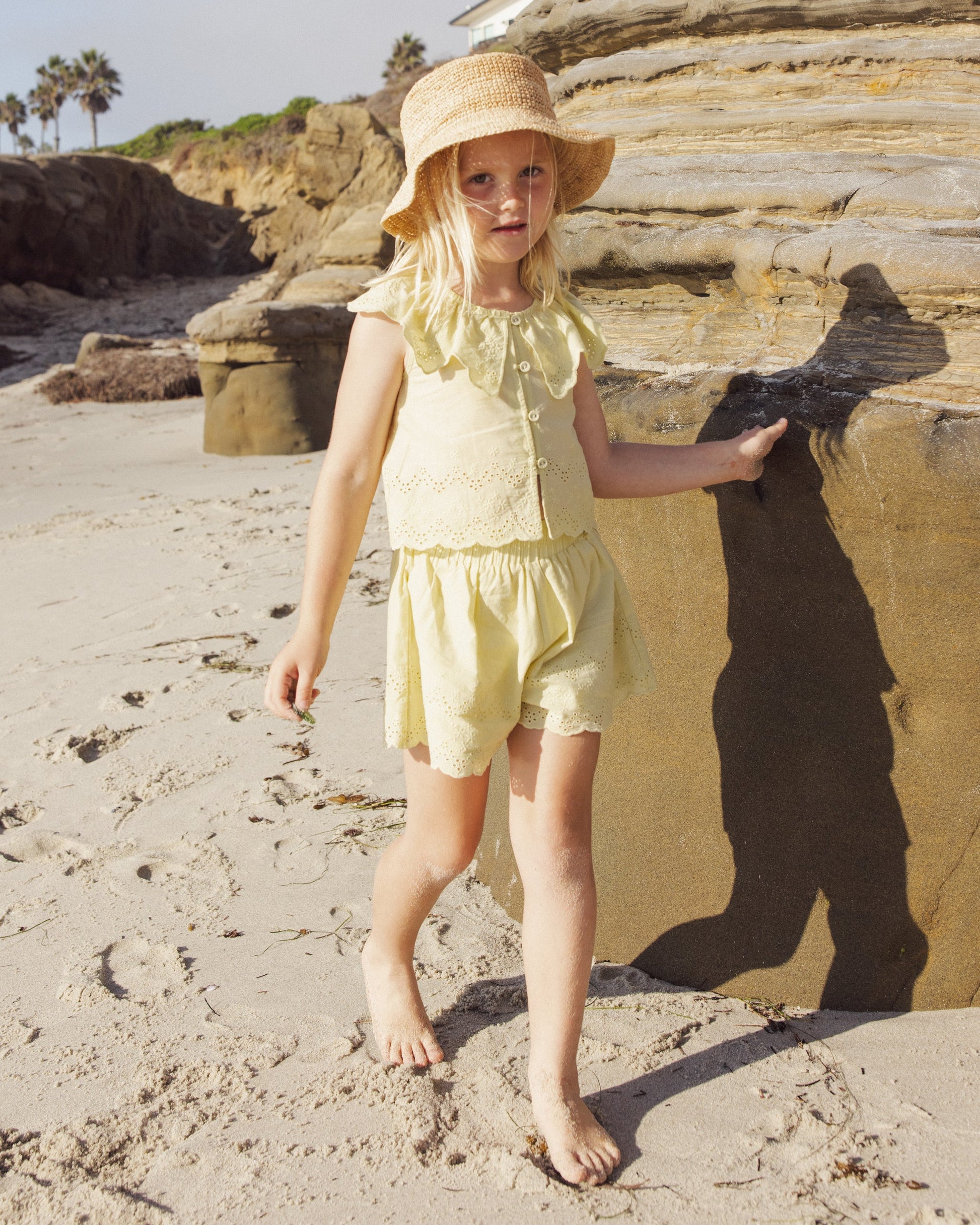A young child in a yellow outfit and sun hat walks barefoot on a sandy beach near large rocks, wearing comfy Rylee & Cru Girls' Remi Shorts with an elastic waist.